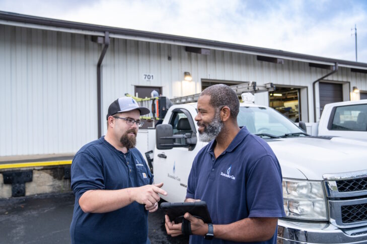 Two men standing in a parking lot talking with a work truck in the background behind them.